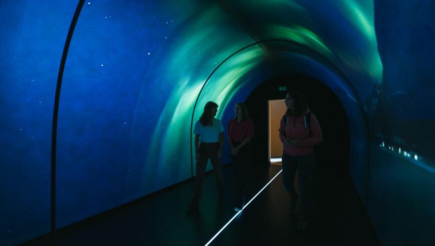 Tre personer går gjennom en tunnel opplyst i nordlysfarger.