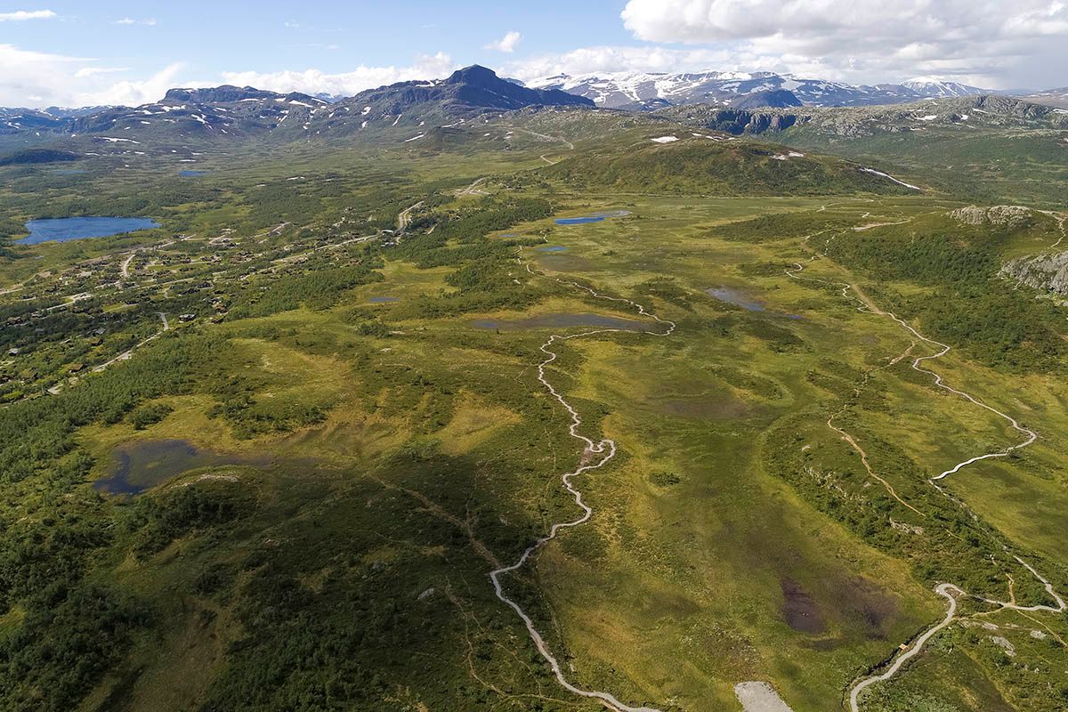Luftfoto av en fjellslette med en grussti for terrengsyklister rundt et myr med høye fjell i bakgrunnen.