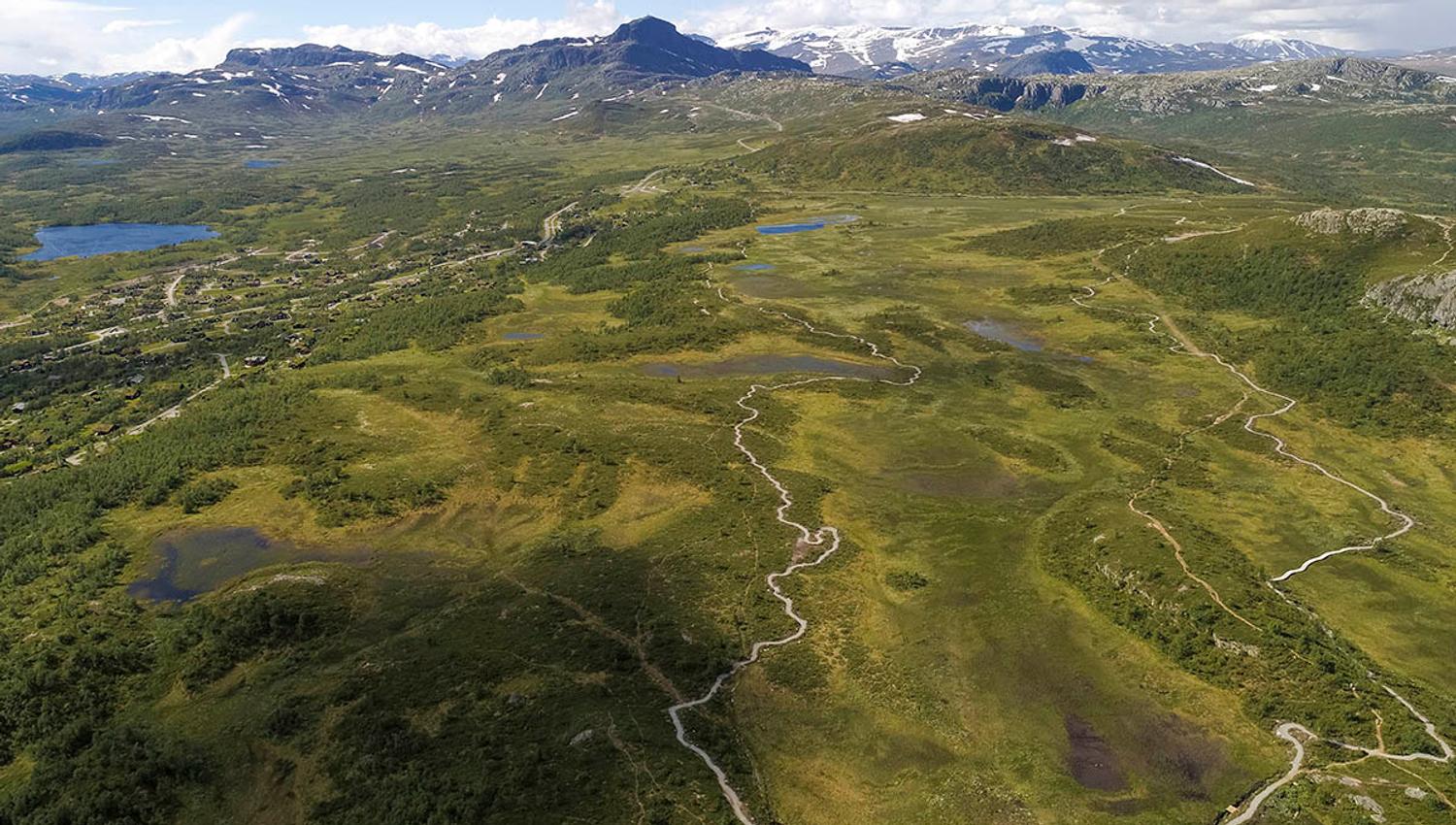 Luftfoto av en fjellslette med en grussti for terrengsyklister rundt et myr med høye fjell i bakgrunnen.