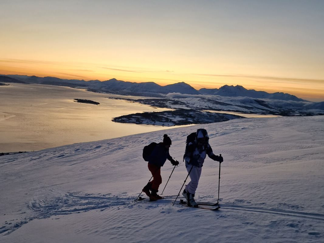 Two guests ski calmly through the snow, while the fjords reflect the late afternoon sun