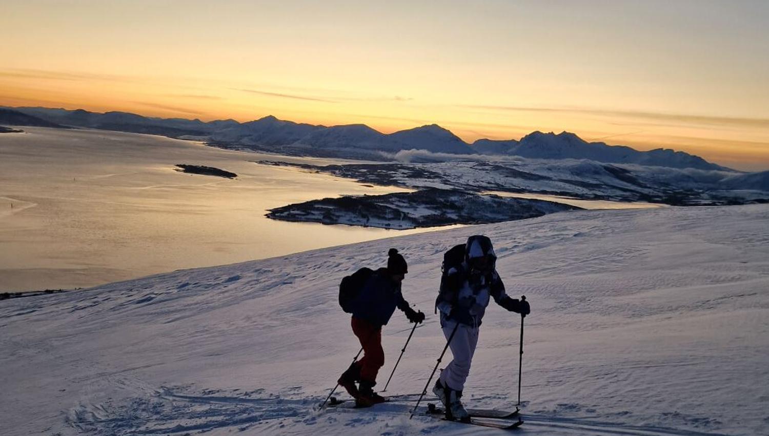 Two guests ski calmly through the snow, while the fjords reflect the late afternoon sun