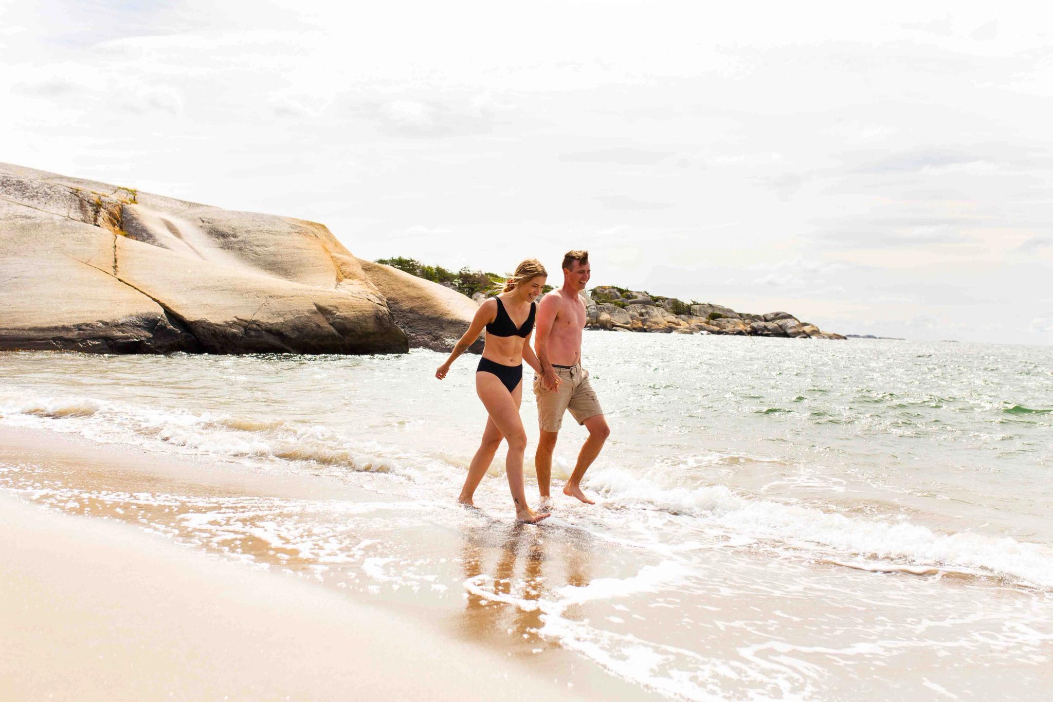 To mennesker går på sandstrand ved svaberg