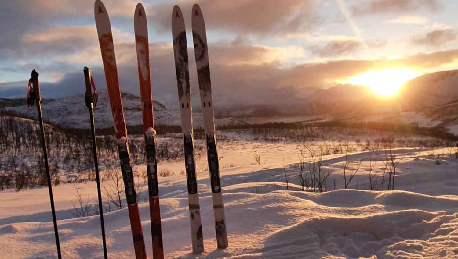 Skis and poles sit ready in the snow while the sun shines bright behind
