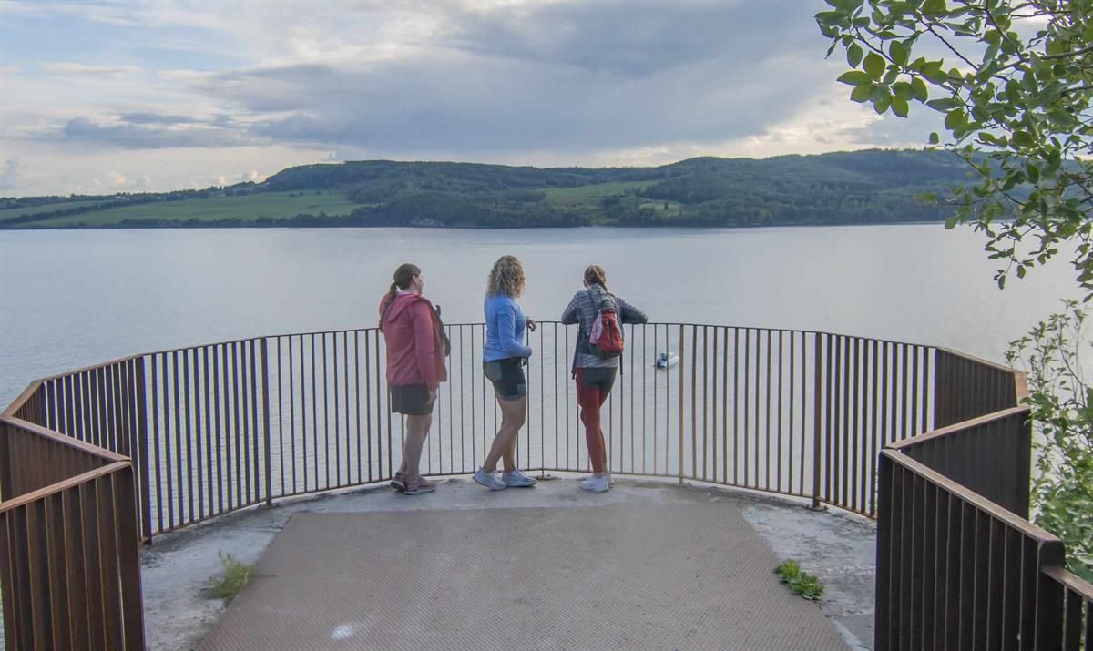 Tre personer står på en utsiktsplattform ved en innsjø og ser utover vannet.