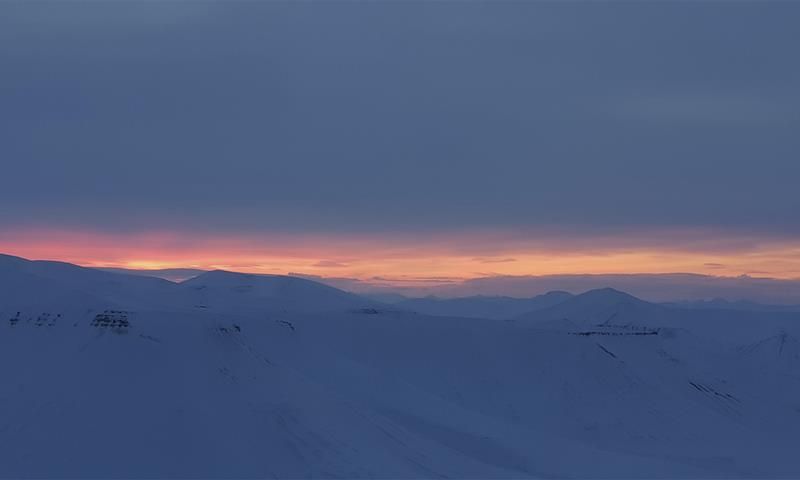 Et snødekt landskap med lave skyer og en tynn stripe med lys fra en solnedgang fjernt i bakgrunnen
