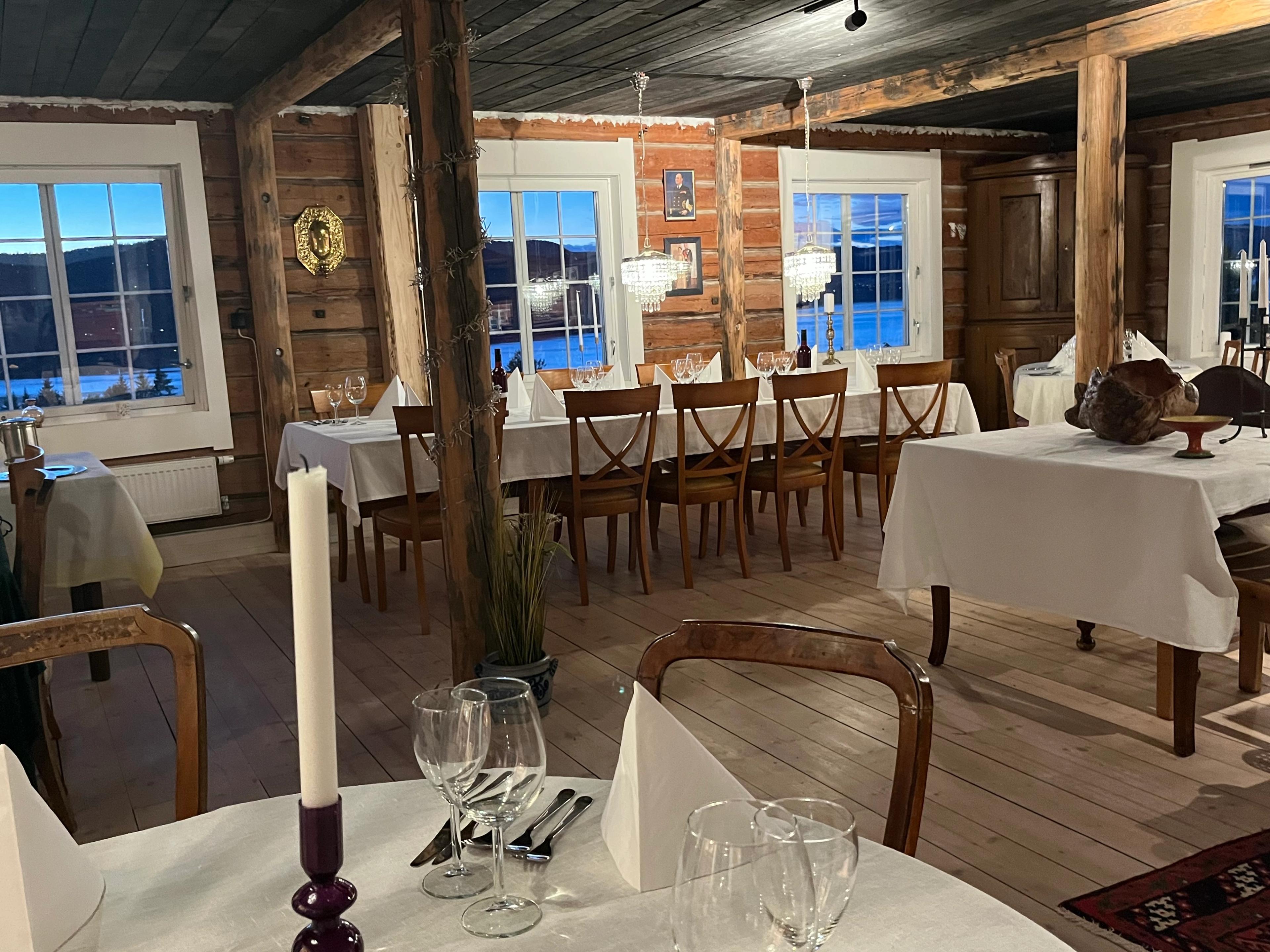 Lys spisestue med trevegger, store vinduer og bord dekket med hvite duker, lysestaker og vinglass