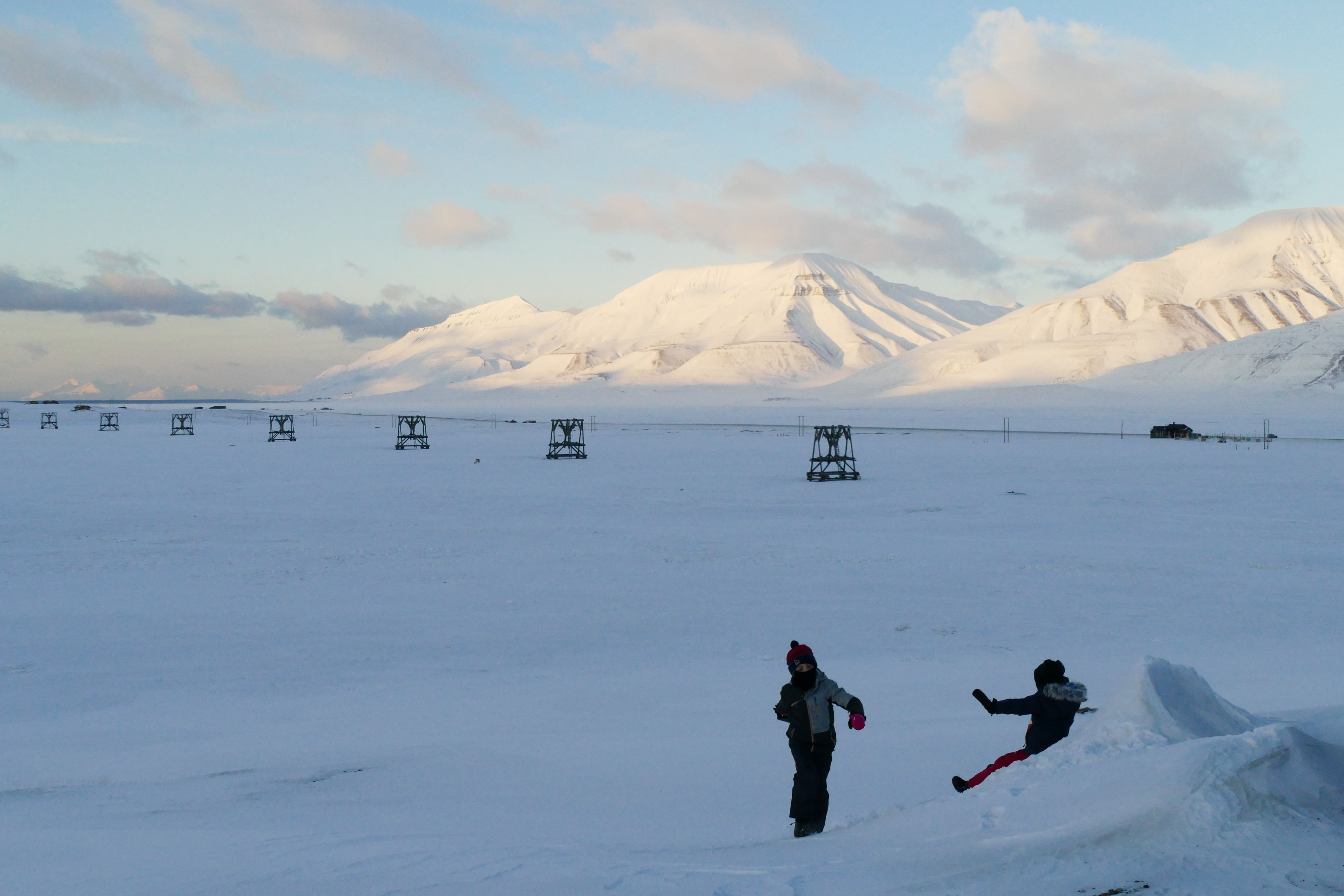 A small person walking up a small hill and another small person sliding down the snowy hill. With historic buildings and snow-covered mountains in the