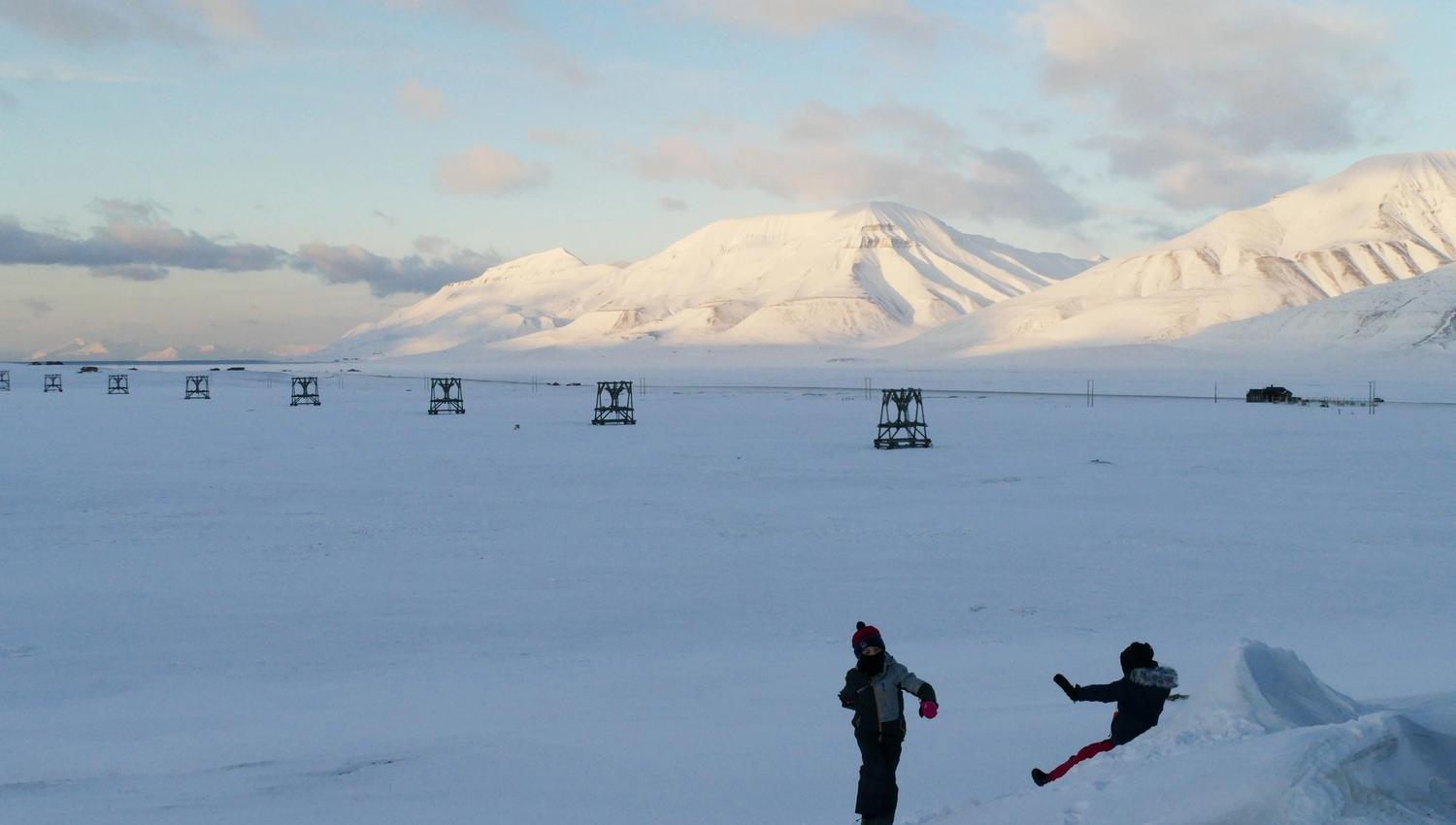 A small person walking up a small hill and another small person sliding down the snowy hill. With historic buildings and snow-covered mountains in the