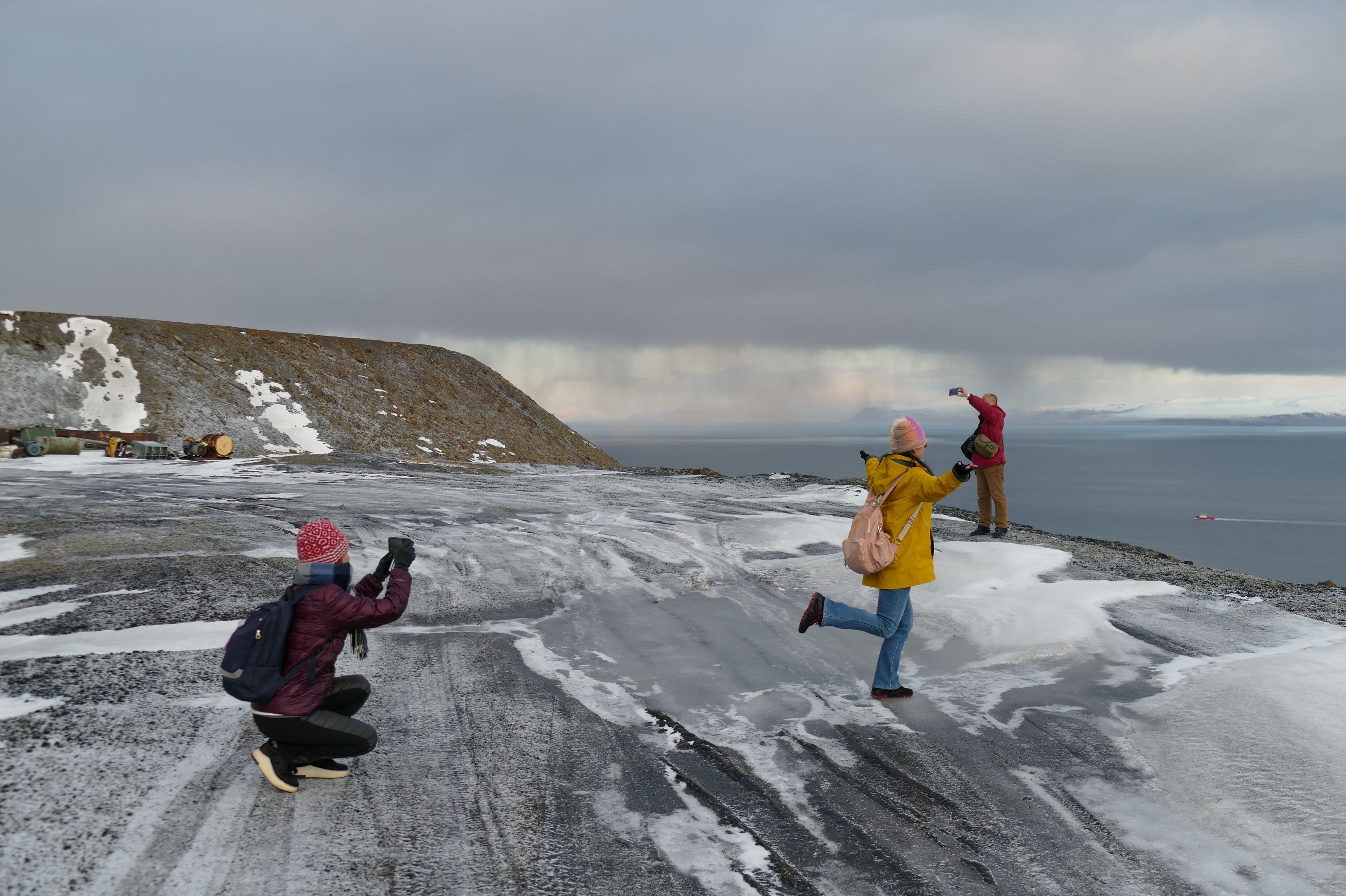 Three people posing to take a photo on snowcovered grounds with a mountain and the ocean in the background