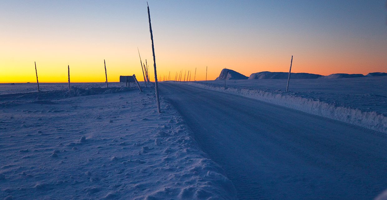 Brøytet vei på førvinterstid etter solnedgang i begynnende blåtime, mens solen restlys fargelegger horisonten gul-oransje. Brøytestikker og en topp horisonten stikker opp fra snøen.