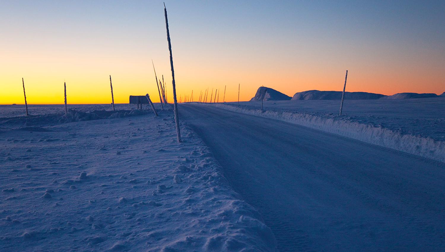 Brøytet vei på førvinterstid etter solnedgang i begynnende blåtime, mens solen restlys fargelegger horisonten gul-oransje. Brøytestikker og en topp horisonten stikker opp fra snøen.
