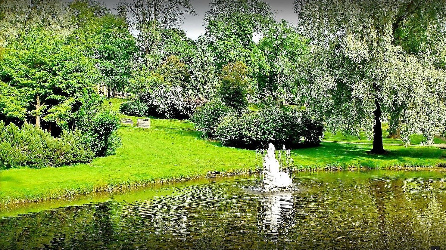Grønn park med trær og busker rundt en dam, der en liten fontene spruter vann og lager ringer i overflaten..