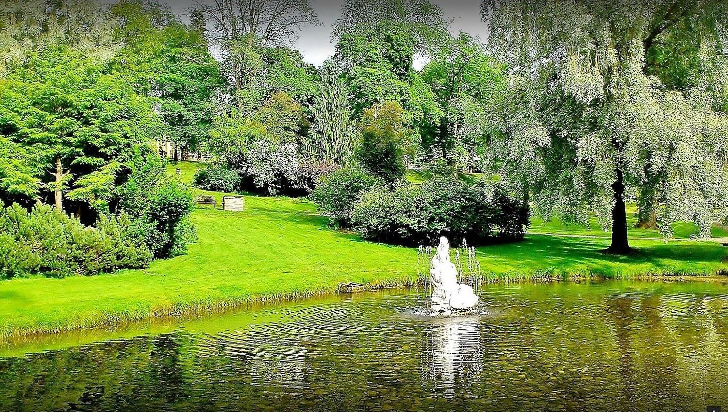 Grønn park med trær og busker rundt en dam, der en liten fontene spruter vann og lager ringer i overflaten..