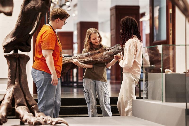 Tre barn undersøker et stort fossil i et museum, mens en voksen hjelper og forklarer.