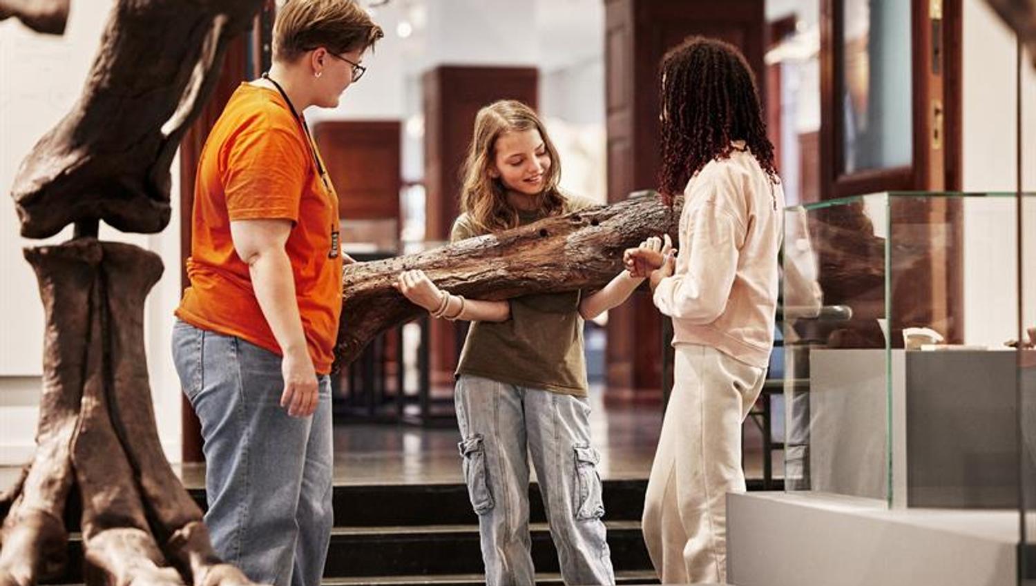 Tre barn undersøker et stort fossil i et museum, mens en voksen hjelper og forklarer.