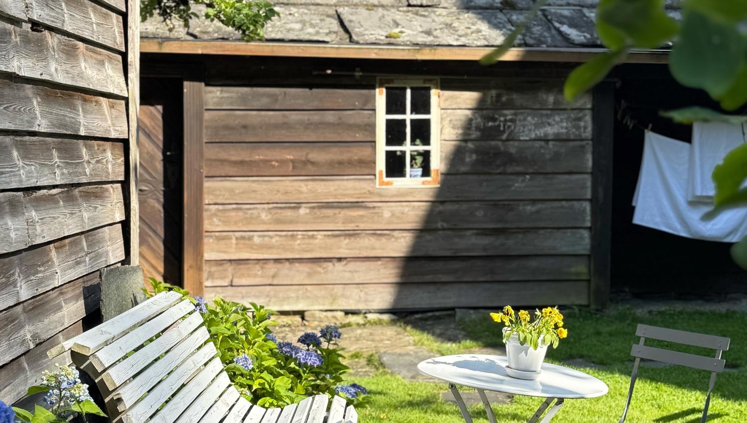 Ein liten hageplass i Agatunet med kvitmåla benk, eit lite bord med blomar og ein stol, omgjeve av blå hortensia framfor eit gamalt tømmerhus.