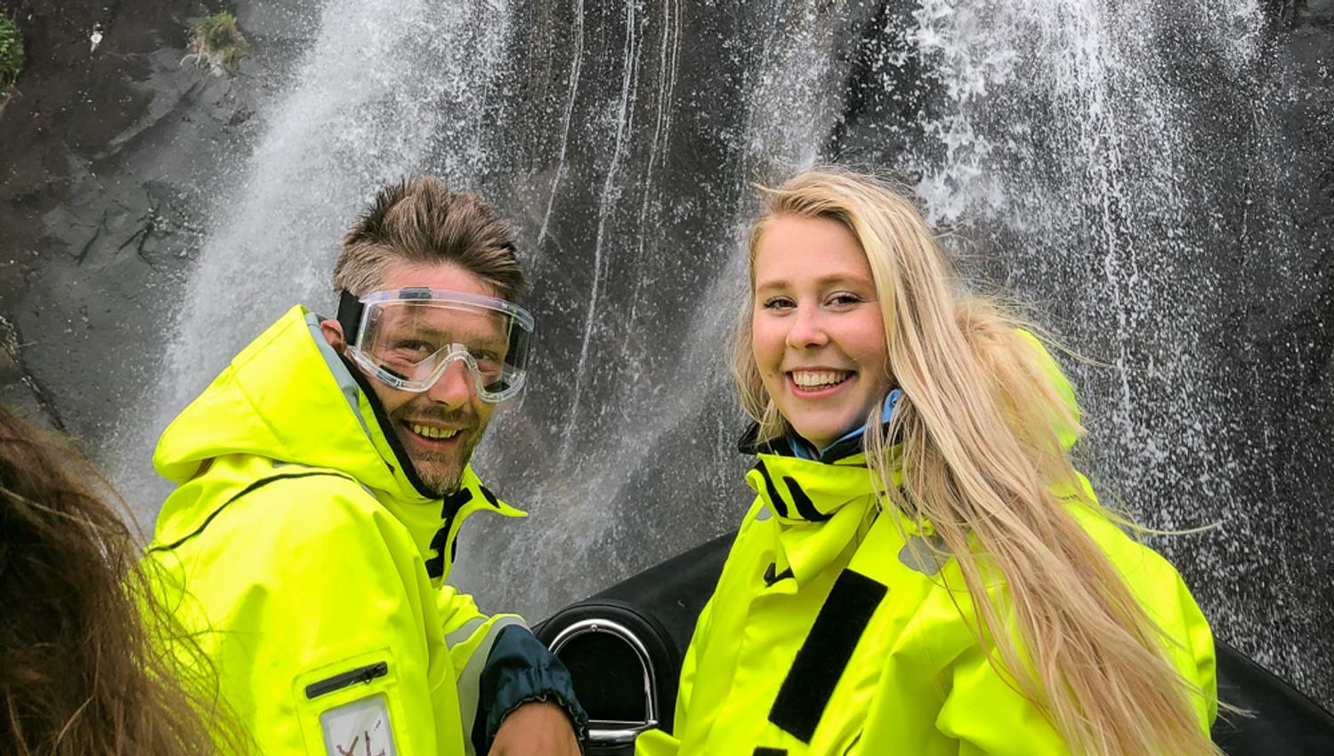 Smilande passasjerar i gule redningsdrakter nyt ei frisk oppleving under ein foss på RIB-safari i Hardanger.