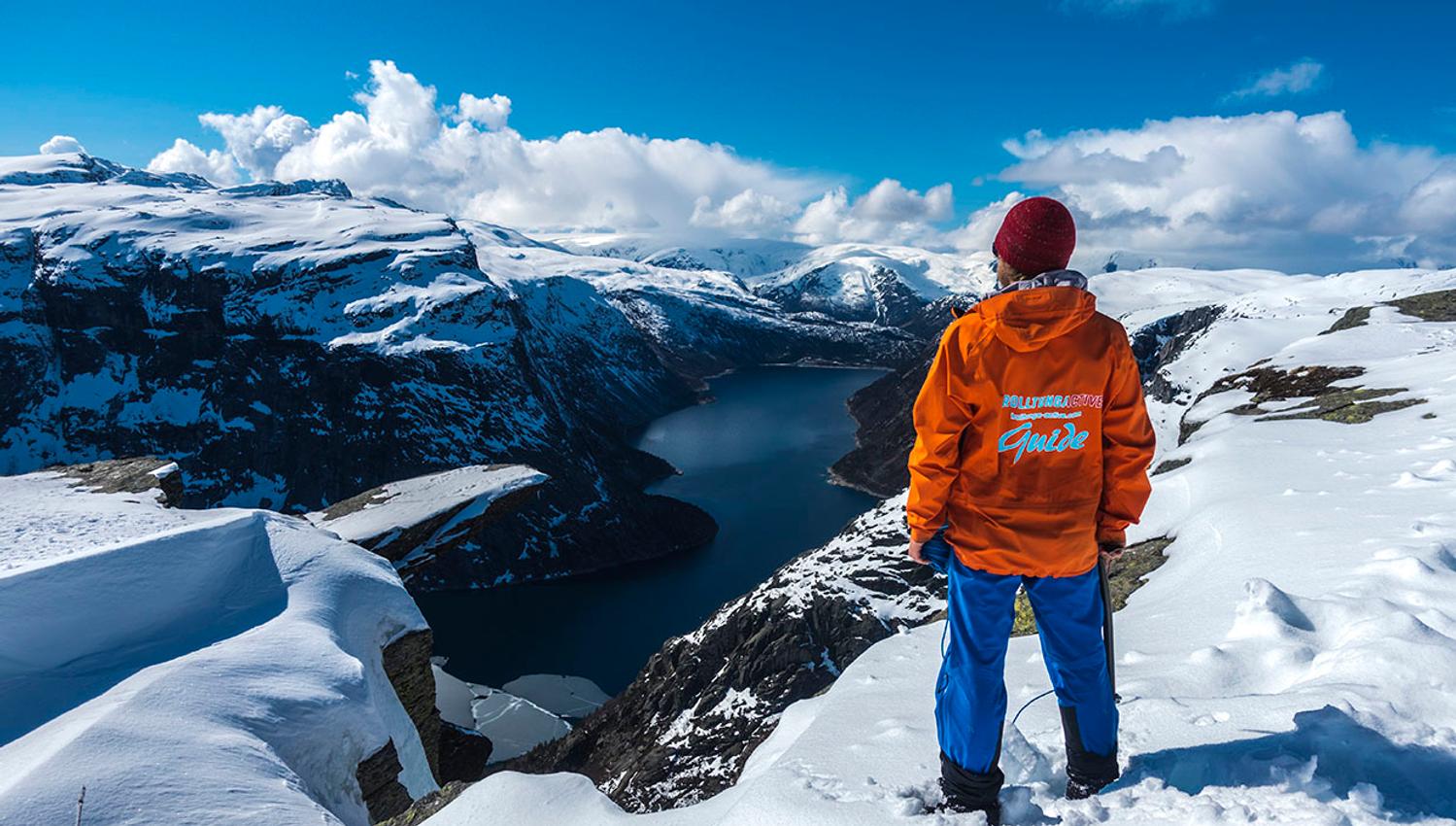 Guide in an orange jacket overlooking the fjord and winter landscape at Trolltunga.