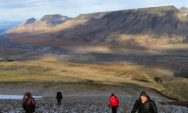 Personer som går opp en bratt bakke med en liten elv og store fjell i bakgrunnen