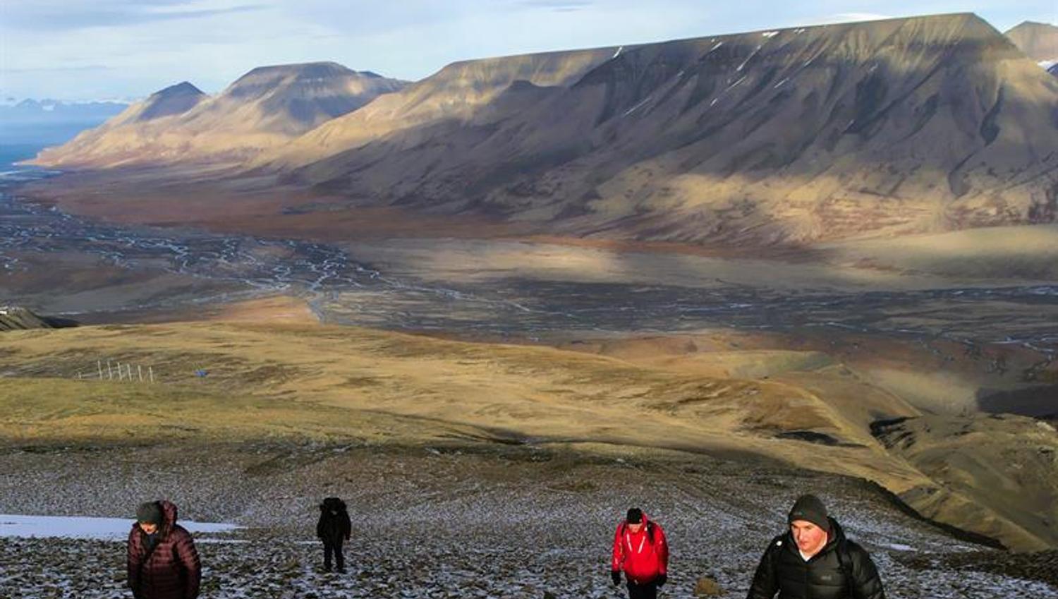Personer som går opp en bratt bakke med en liten elv og store fjell i bakgrunnen