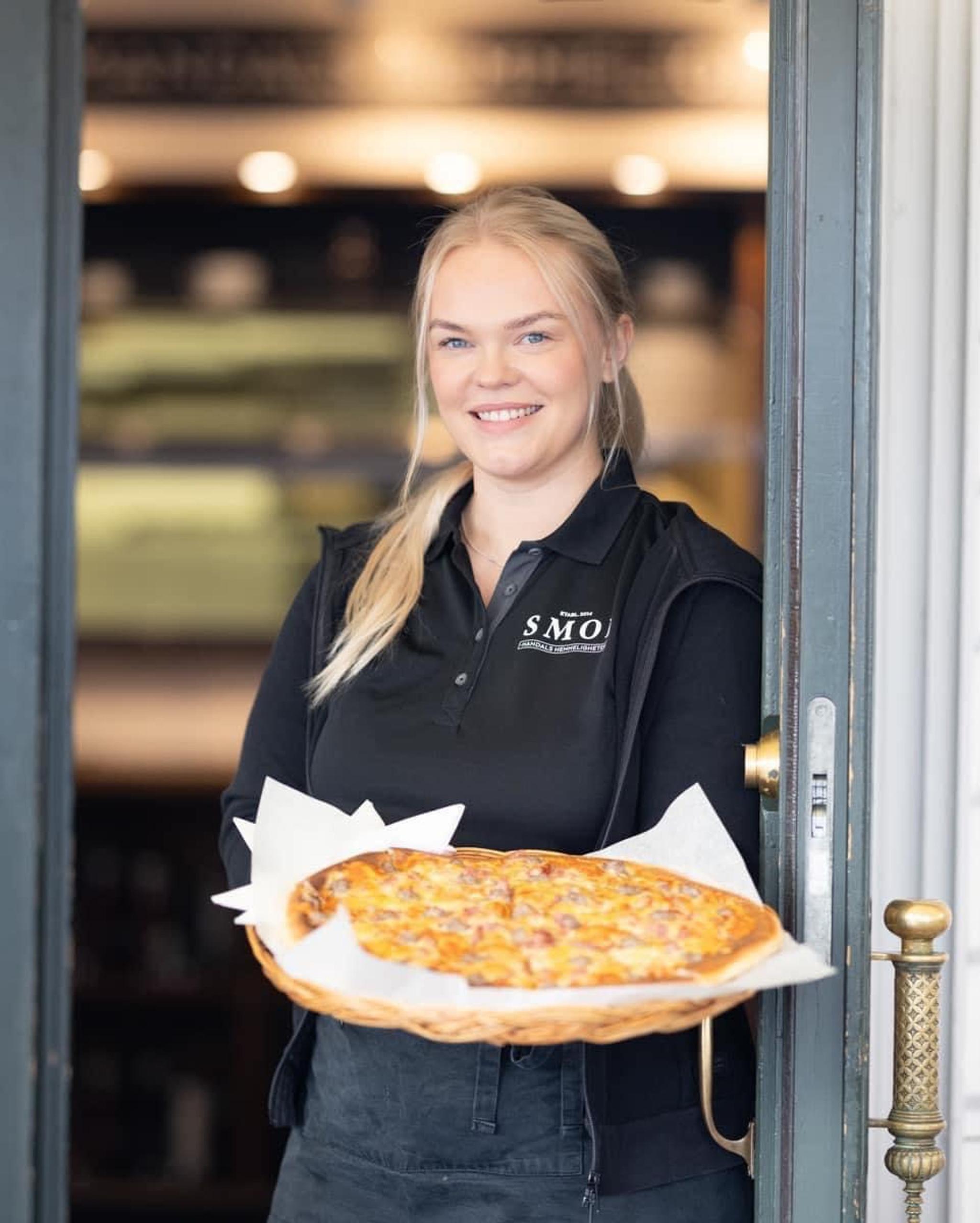 Blid servitør fra SMOI som står i døra og holder en deilig pizza