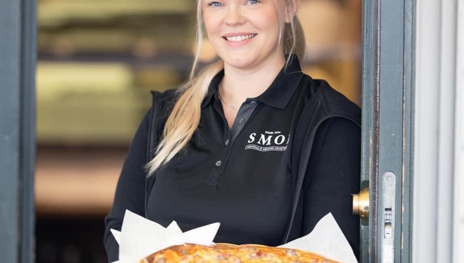 Blid servitør fra SMOI som står i døra og holder en deilig pizza
