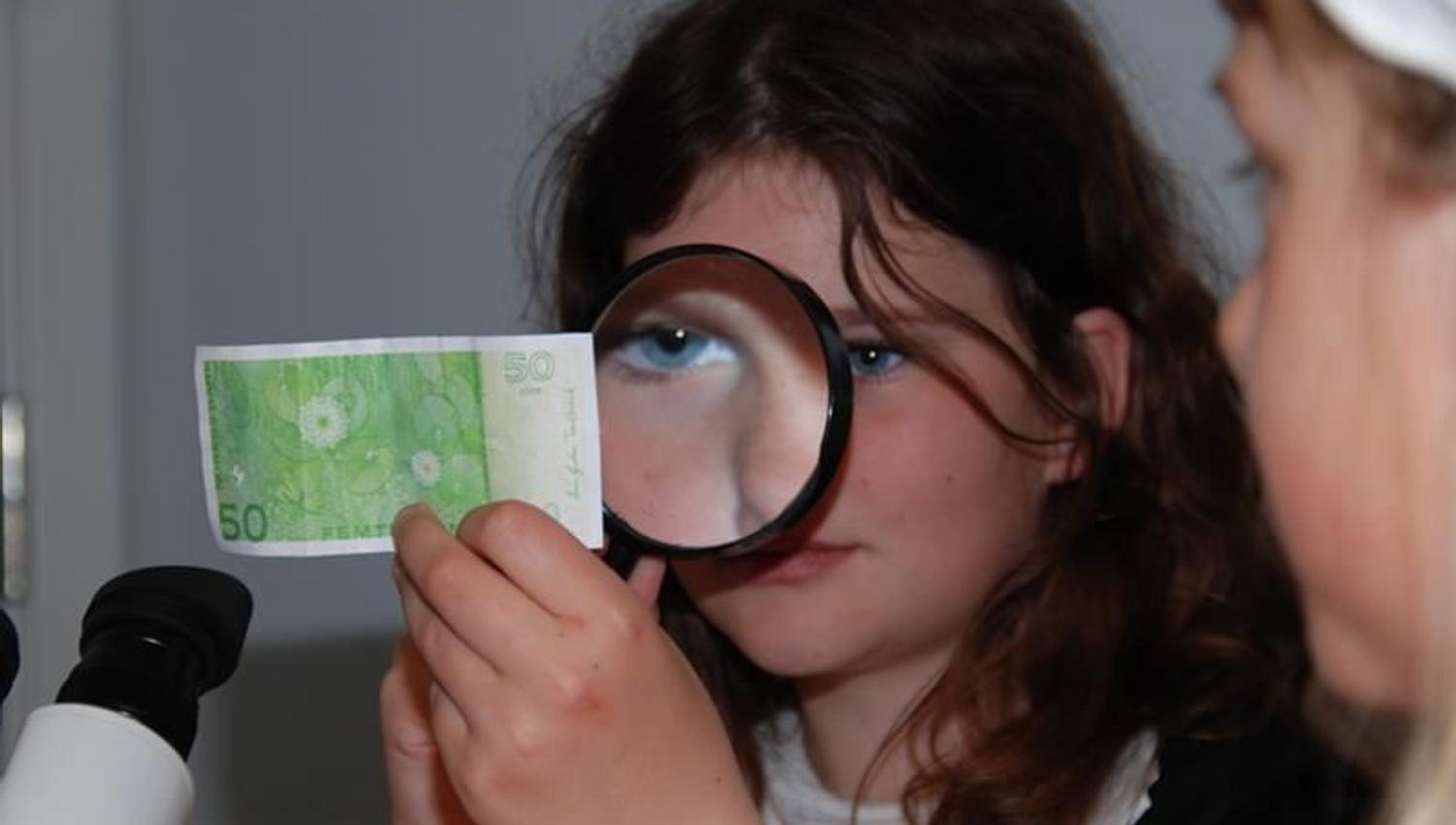 A girl look closer at a 50 kroner note with a magnifying glass