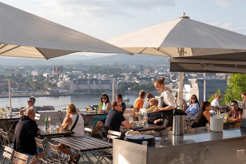 A waiter serves food to guests on a lively rooftop terrace overlooking the city.