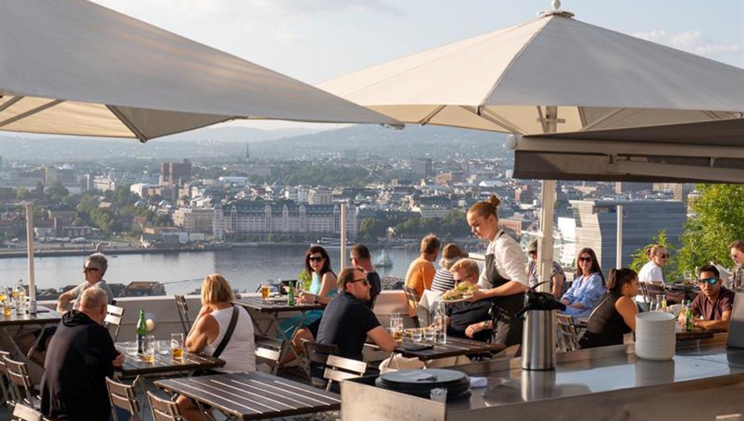 A waiter serves food to guests on a lively rooftop terrace overlooking the city.
