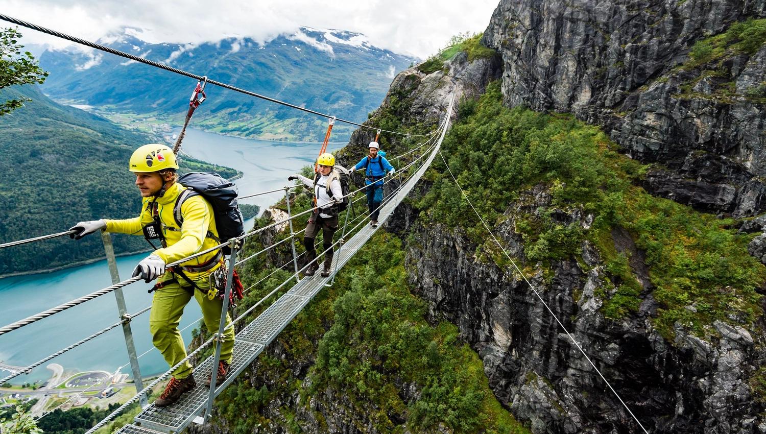 Tre klatrarar med hjelm og sele kryssar ei smal hengebru langs ei bratt fjellside over Nordfjord.