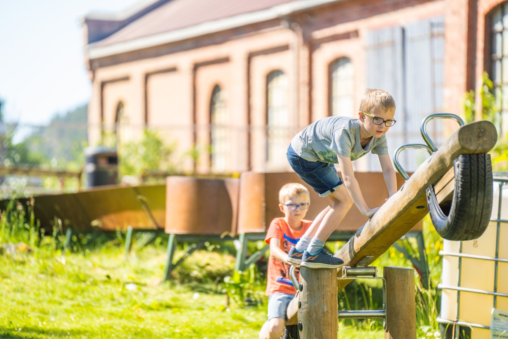 Bildet viser to gutter som leker ute på en trevippe laget av en stokk og et dekk. Den ene gutten klatrer opp mens den andre står bak og ser på. Det er sol og grønt gress, og i bakgrunnen står en stor murbygning.