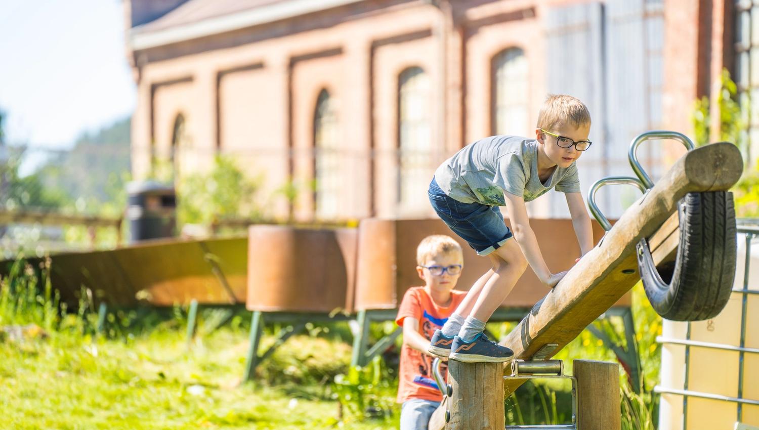 Bildet viser to gutter som leker ute på en trevippe laget av en stokk og et dekk. Den ene gutten klatrer opp mens den andre står bak og ser på. Det er sol og grønt gress, og i bakgrunnen står en stor murbygning.