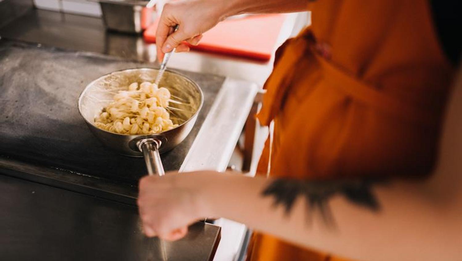 A person wearing an orange apron is stirring a small pot of macaroni and cheese