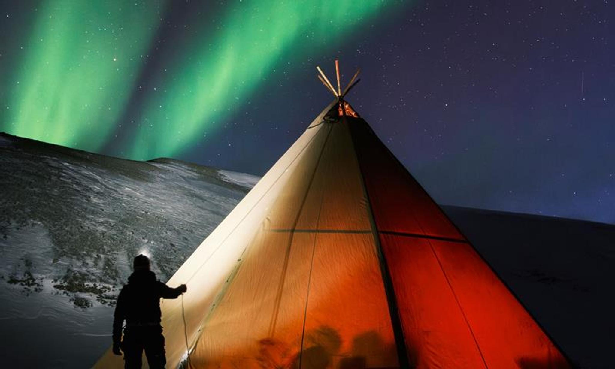 A lighted tent. A person standing outside with headlamp. Green northern ligths on the sky.
