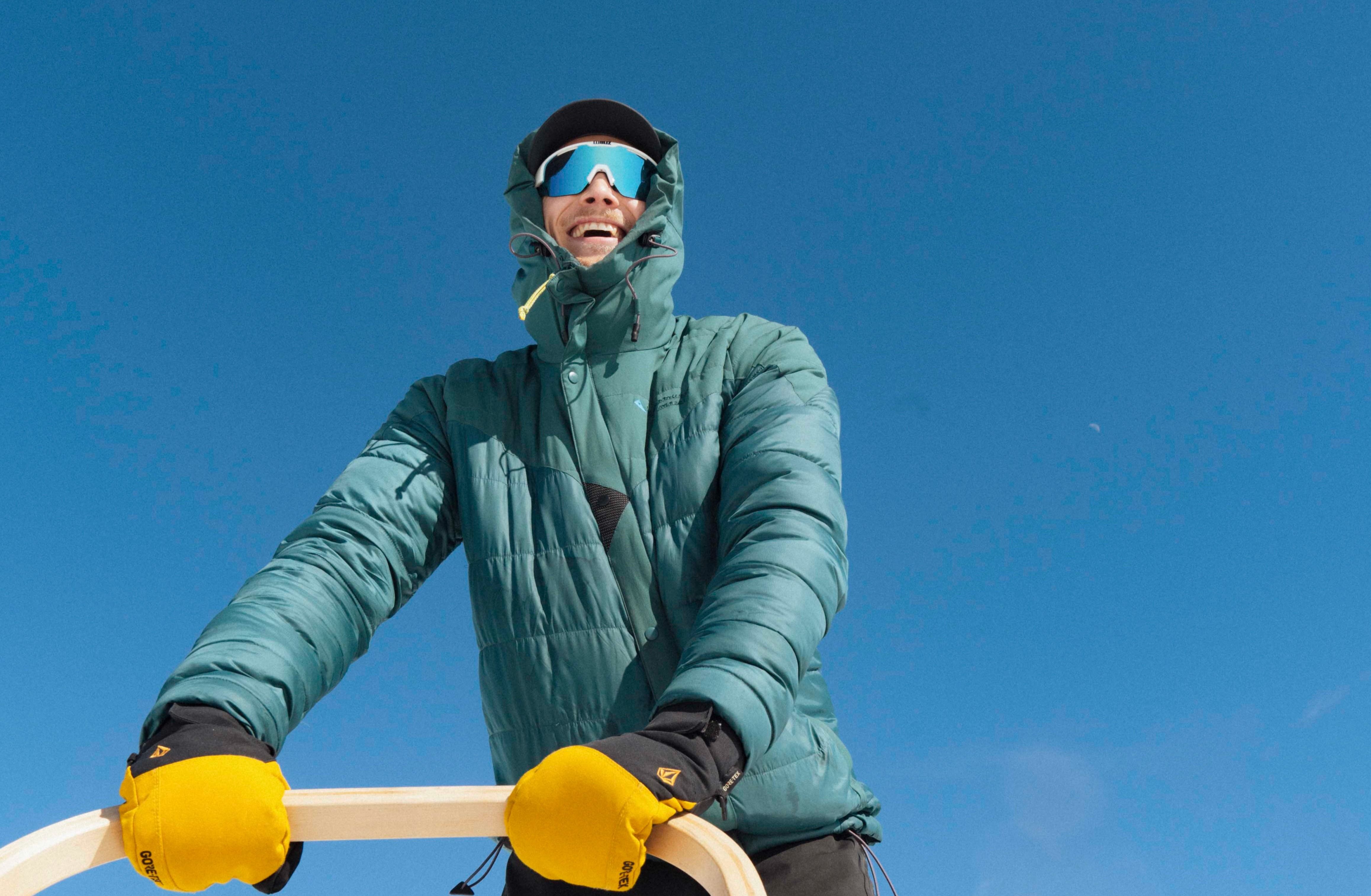 En person kledd i varm jakke og gule hansker står bak et hundespann med huskier på folgefonna husky camp