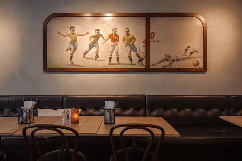 Classic restaurant interior with upholstered bench seating, wooden tables and a wall mural of football players.