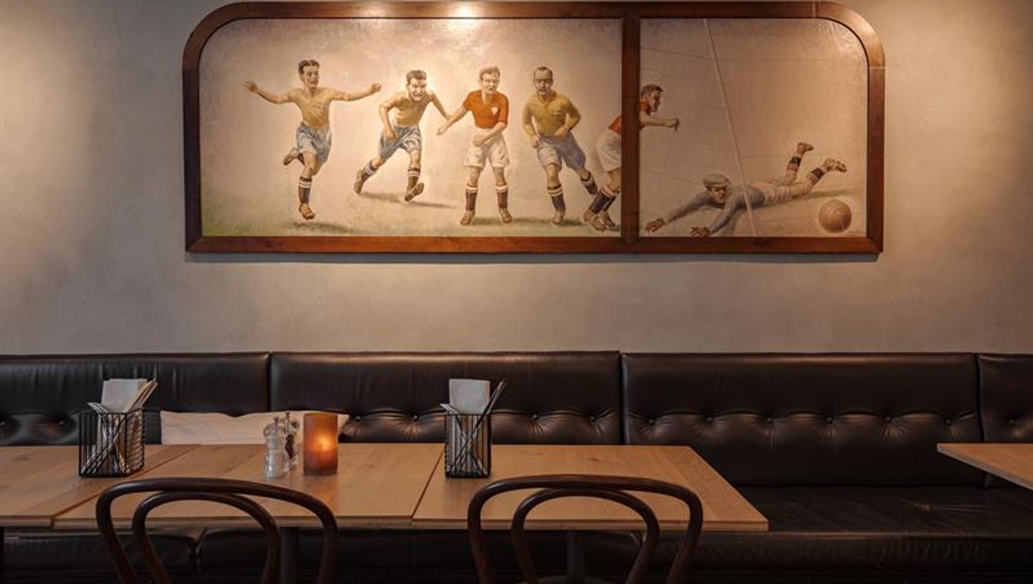 Classic restaurant interior with upholstered bench seating, wooden tables and a wall mural of football players.