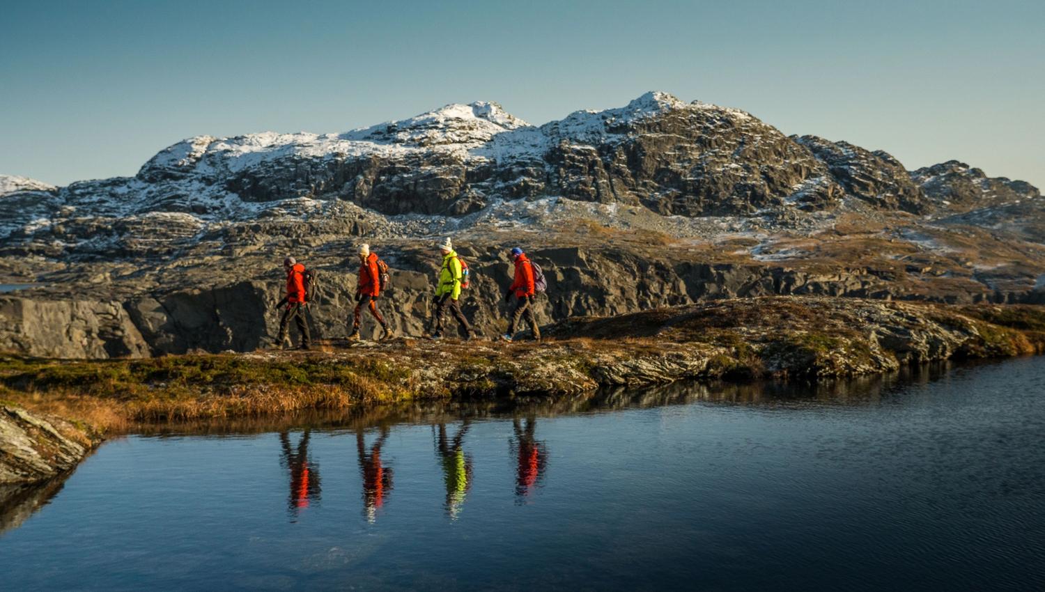 Group of hikers walking by a calm mountain lake with snow-covered peaks near Trolltunga.
