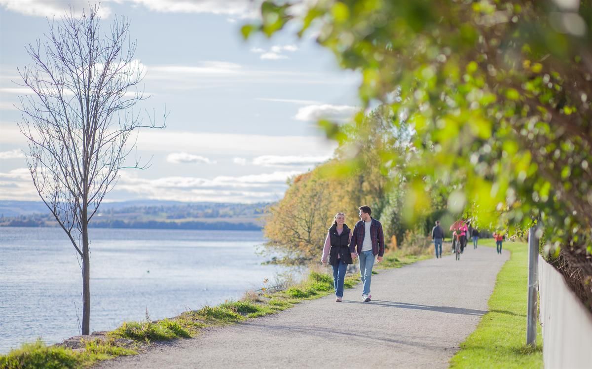 Folk går langs en gangvei ved vannet på en lys dag med trær på siden.