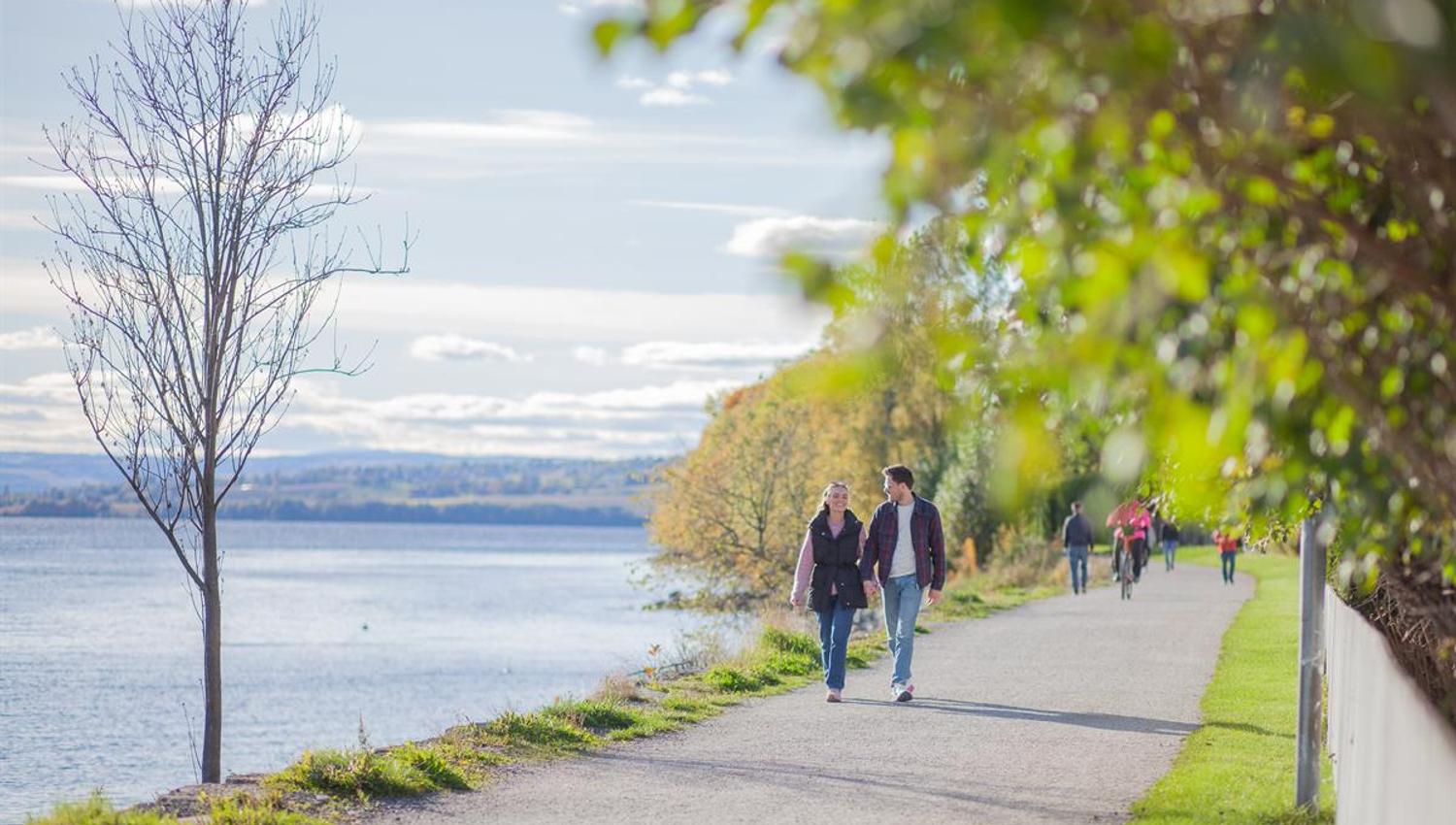 Folk går langs en gangvei ved vannet på en lys dag med trær på siden.