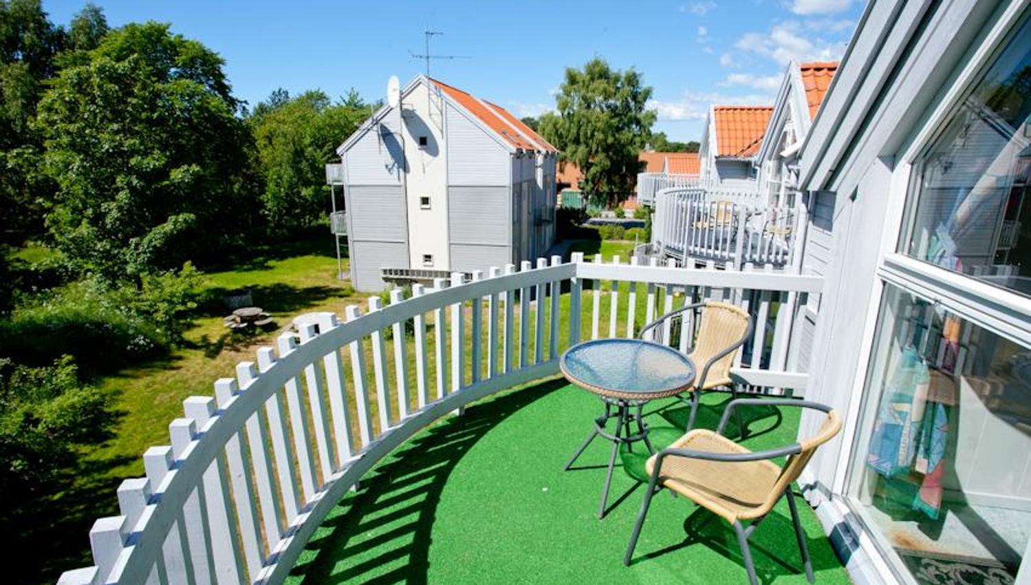 Liten balkong med hvitt gjerde og kunstgress. to nettingstoler og lite bord.