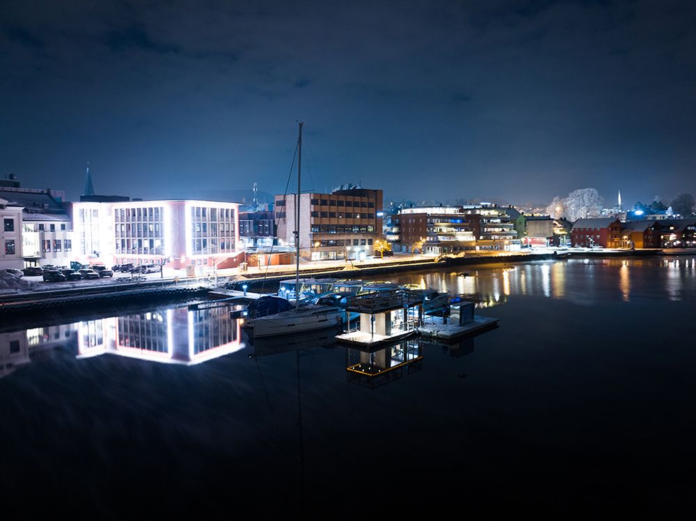 sauna liggene ved elva, med båter på brygge. blokker med lys i bakgrunnen