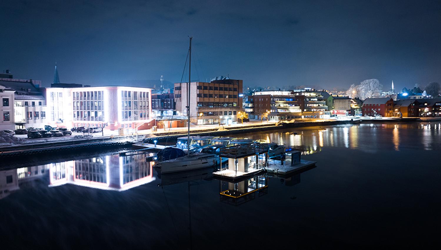 sauna liggene ved elva, med båter på brygge. blokker med lys i bakgrunnen