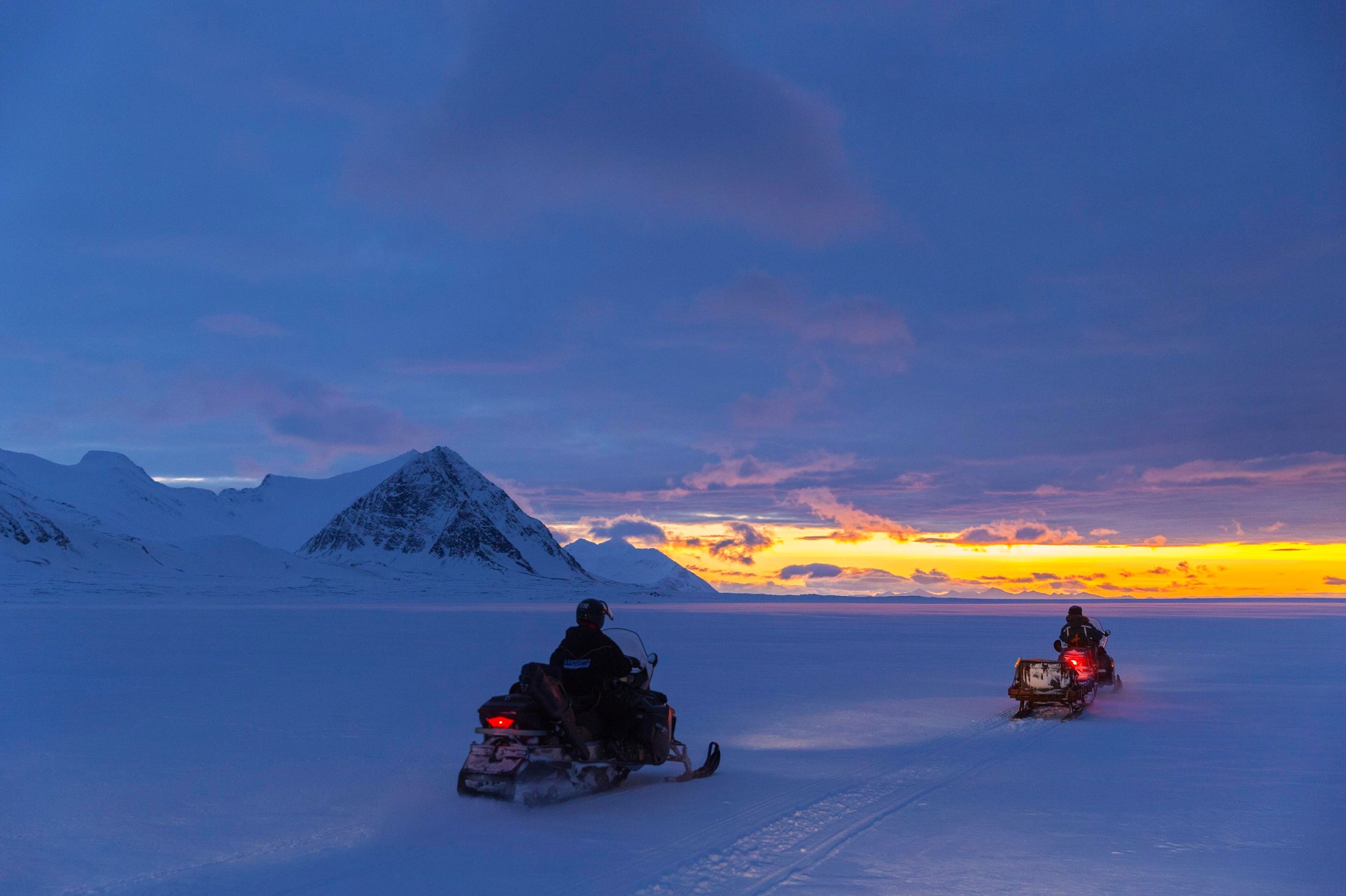 A guest and a guide driving snowmobiles at dusk. In the background there's sunlight on the horizon. 