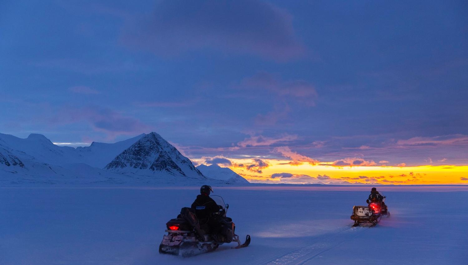 A guest and a guide driving snowmobiles at dusk. In the background there's sunlight on the horizon.