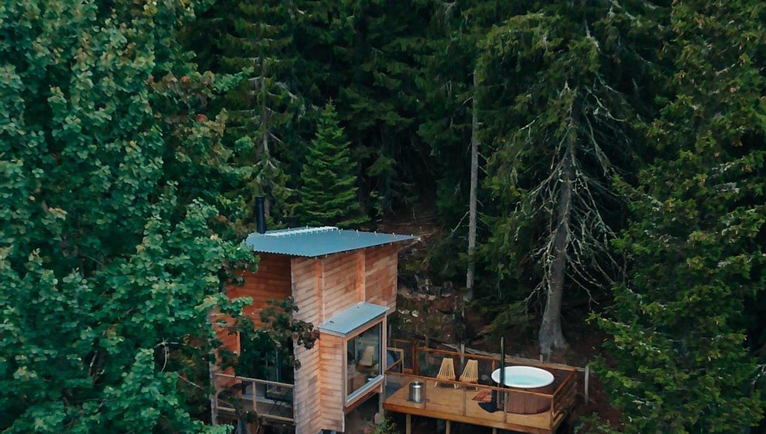 Wooden cabin on stilts surrounded by dense green forest, viewed from above.