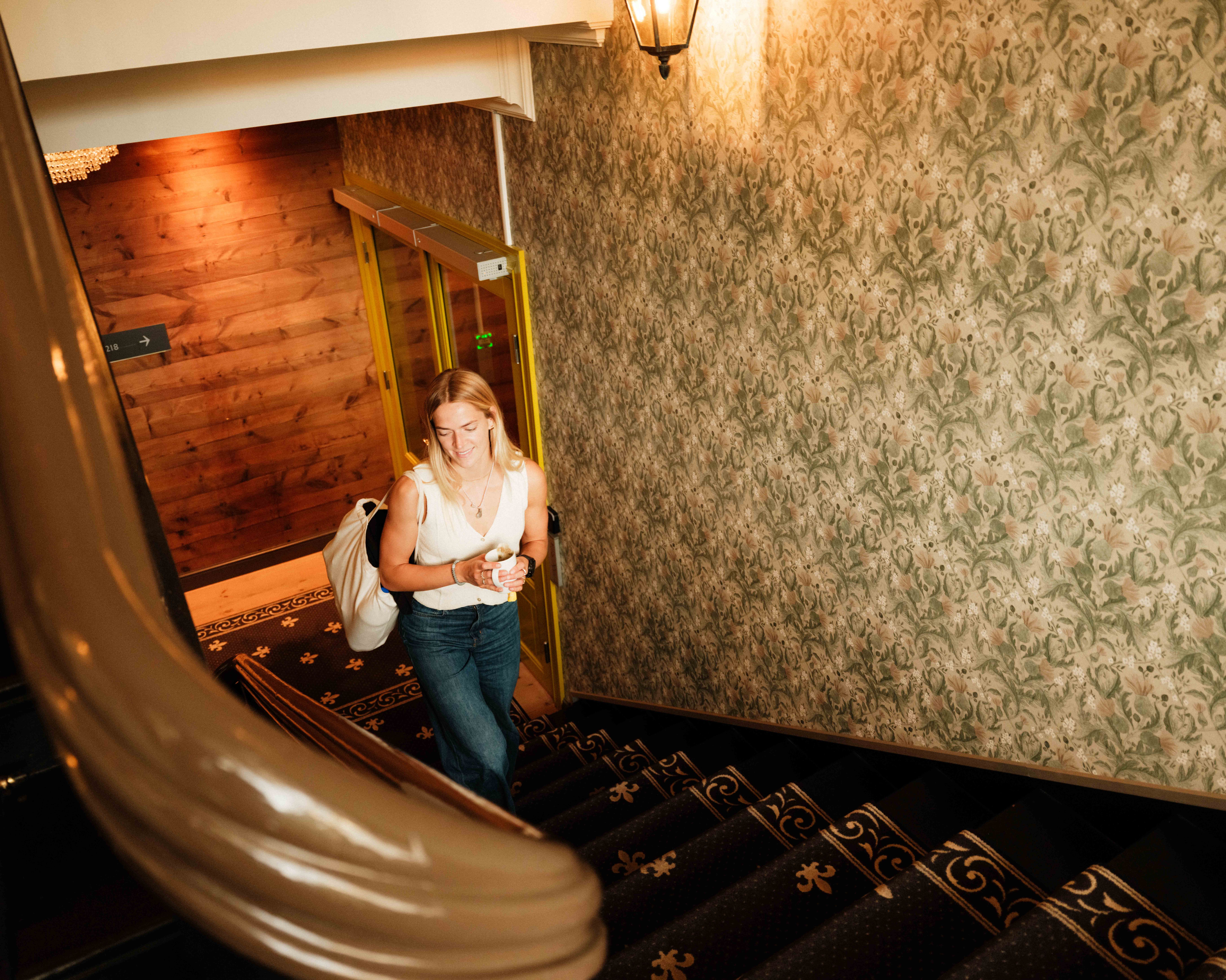 Ei gjest går opp den originale trappa i Thon Hotel Sandven – med kaffikopp og bagasje i handa.