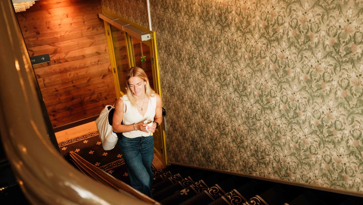 Ei gjest går opp den originale trappa i Thon Hotel Sandven – med kaffikopp og bagasje i handa.