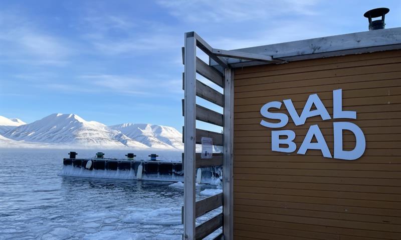 SvalBad sin logo på utsiden av badstuen med en fjord og snødekte fjell i bakgrunnen.