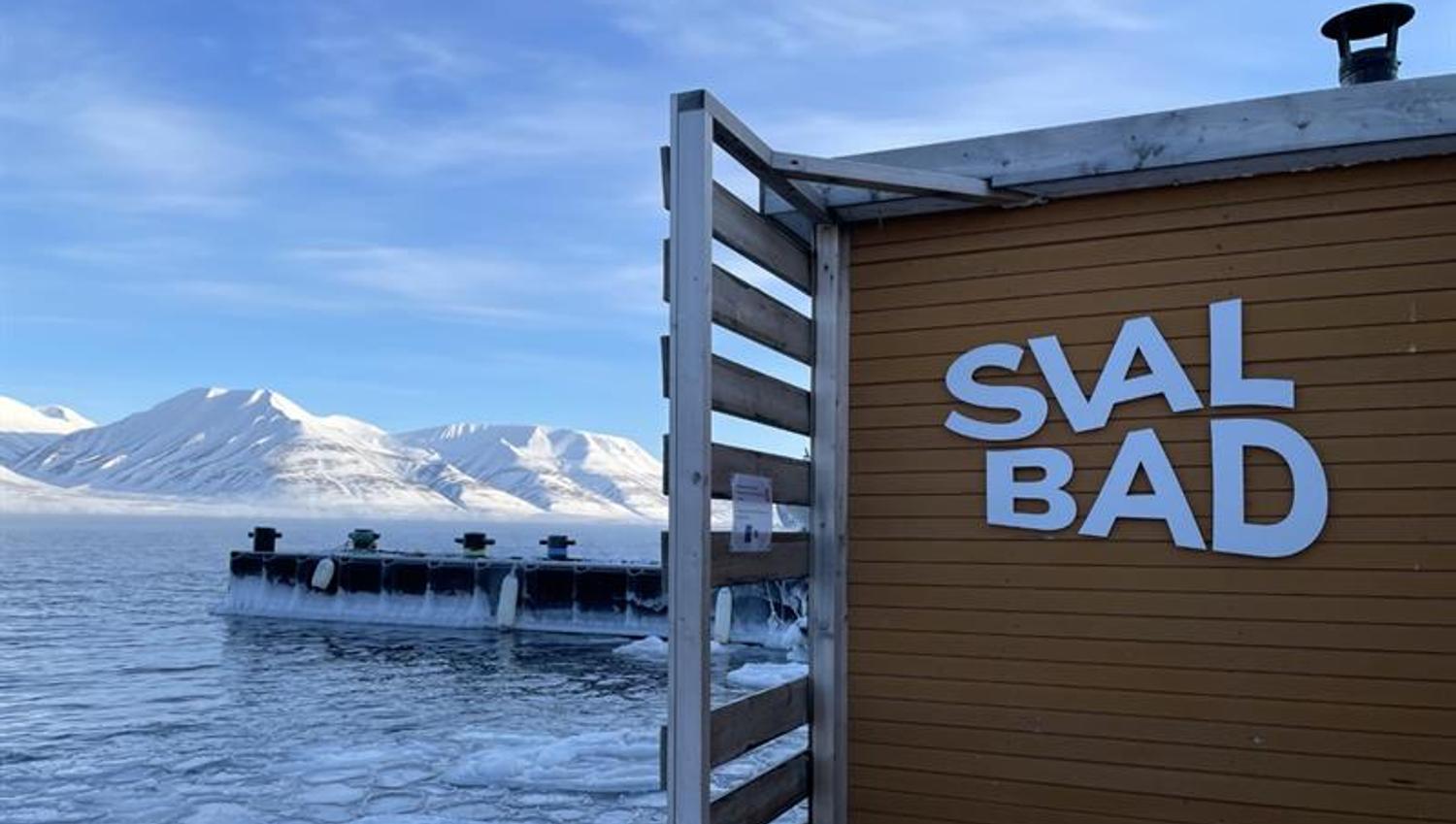 SvalBad sin logo på utsiden av badstuen med en fjord og snødekte fjell i bakgrunnen.